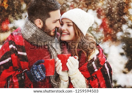 Young beautiful happy smiling couple holding red cups, mugs of hot drink. Models looking at each other, bundled up tartan blanket. Winter holidays, Christmas celebration, winter holidays concept
