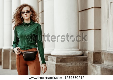 Outdoor fashion portrait of young lady wearing sunglasses, green turtleneck, brown trousers, leather belt bag, necklace, bracelet, posing in street of european city. Copy, empty space for text