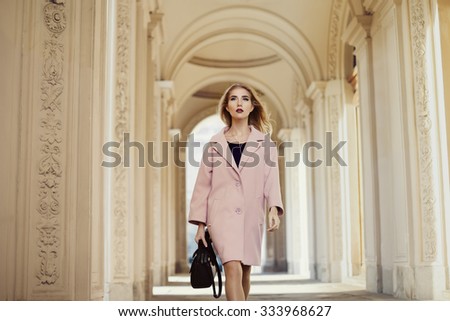 Street fashion concept: portrait of young beautiful woman  wearing pink coat with handbag walking on old architecture background