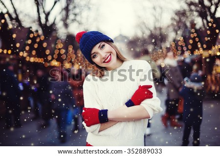 Street portrait of smiling beautiful young woman on the festive Christmas fair. Lady wearing classic stylish winter knitted clothes. Model looking at camera. Magic snowfall effect. Close up