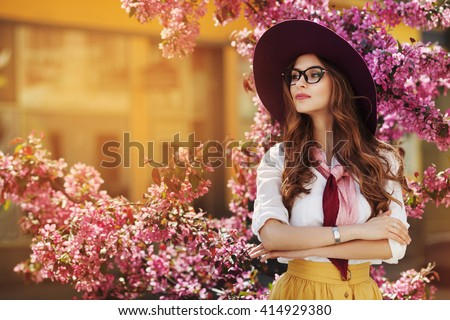 Outdoor portrait of young beautiful fashionable lady posing near flowering tree. Model wearing stylish accessories & clothes. Girl looking aside. Female beauty & fashion. City lifestyle. Copy space
