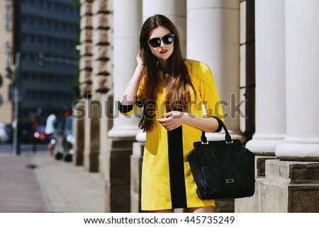 Outdoor portrait of young beautiful lady walking on the street. Model wearing sunglasses & stylish yellow summer dress. Girl looking down. Female fashion concept. City lifestyle. Sunny day. Waist up