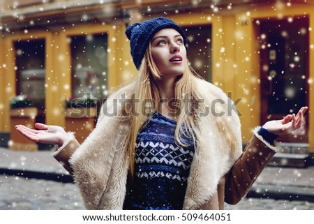 Outdoor portrait of beautiful happy smiling young woman posing on street. Model looking up, at sky. Lady wearing stylish winter clothes. Magic snowfall effect. Christmas, new year concept. Waist up