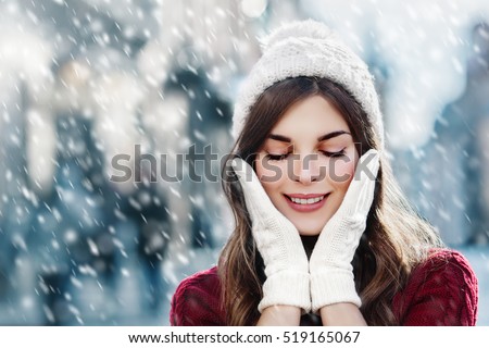 Outdoor close up photo of young beautiful happy smiling girl walking on street. Model closed her eyes and touching face, wearing stylish white knitted winter hat and gloves. Copy, empty space for text