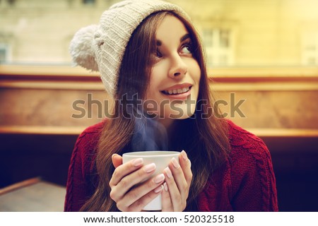 Young beautiful happy smiling girl holding white mug with hot liquid, drinking coffee or tea. Model wearing stylish knitted winter clothes, accessories. Lady looking up. Indoor. Close up. Toned