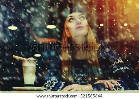 Young beautiful woman sitting in cafe, drinking coffee. Model looking up. Magic snowfall effect. Christmas, new year, winter holidays concept. Waist up. View through the window glass. Toned