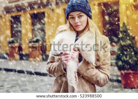 Outdoor waist up portrait of young beautiful fashionable woman posing on street. Model wearing stylish winter fake fur coat and beanie hat, looking at camera.  Magic snowfall effect. Day light
