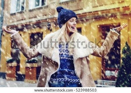 Young beautiful happy smiling girl posing on street. Model spread out her hands, catching snowflakes. Woman wearing stylish winter clothes. Magic snowfall effect Christmas, new year concept. Waist up