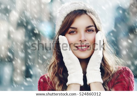 Young beautiful happy smiling girl walking on street. Model looking at camera, touching her face, wearing stylish knitted winter hat and gloves. Close up. Outdoor. Toned. Copy, empty space for text