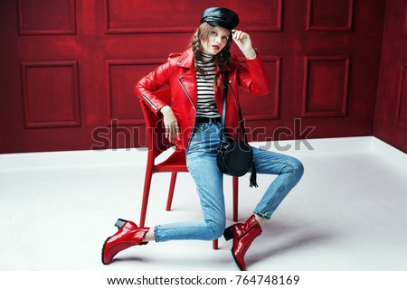 Full body studio fashion portrait of young beautiful woman posing on chair. Model wearing stylish leather cap, biker jacket, stripped turtleneck, blue jeans and red textured ankle boots.