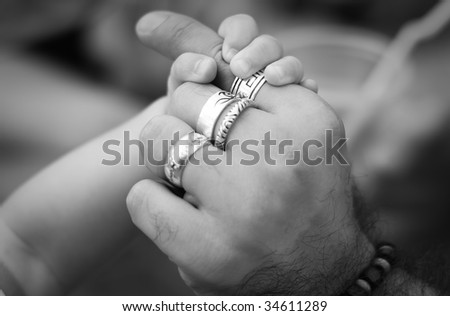 A hand of a baby holding man\'s hand with silver rings and bracelet, black and white
