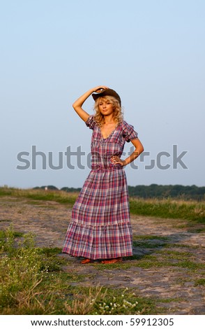 Beautiful young blond woman in a cowboy hat and plaided dress standing on a road in the rural area