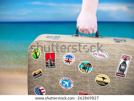 hand holds a suitcase with stickers on background of ocean