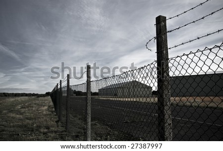 Broken barbed wire fence surrounding an industrial warehouse facility against a dramatic sky