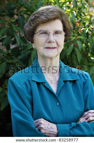 Portrait of mature woman wearing blue blouse with folded arms standing outside in front of flower bush.