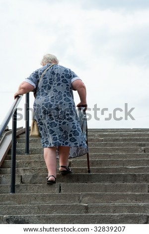 Old grey-haired the woman climbs the stairs by means of a crutch and a handrail