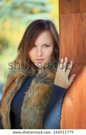 Young beautiful woman in faux fox fur waistcoat standing near wooden column in the park