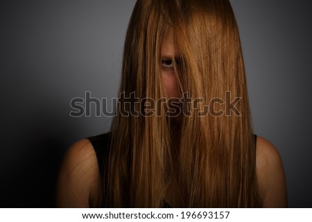 Young woman with long blonde hair standing in dramatic light and angry looking through locks on grey background