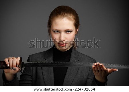 Beautiful young business woman in dark jacket holding japan katana sword on grey background