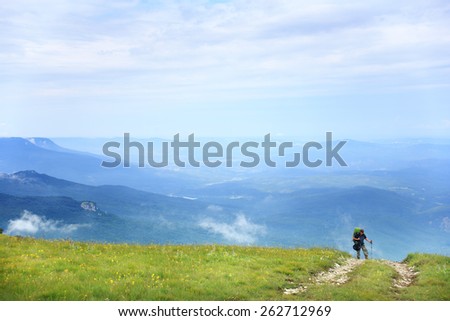 Young woman with a backpack and trekking poles walking on the path up the hill with green grass on the background of the beautiful mountain scenery under the blue summer sky with cloudscape