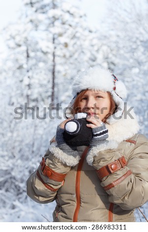 Beautiful young thoughtful woman in down jacket, fingerless mittens and hat with white fur holding knitted camera at the background of snowy forest.