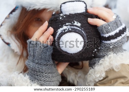 Beautiful young woman in winter clothing, fingerless mittens and hat with white fur photographing by a black knitted camera. Shallow dof. Focus on camera.