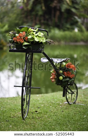 A wrought iron bicycle decorated with flowers on a green grass patch with a pond in the background