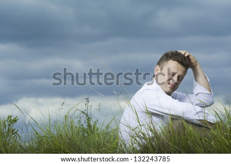 Young businessman sitting on the meadow. Thinking about something and scratching his head. Sky and meadow in the background.