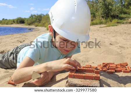 to play in construction. boy in a helmet is building a house out of bricks on the river bank