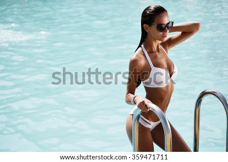 Elegant sexy woman in the white bikini on the sun-tanned slim and shapely body is posing near the swimming pool