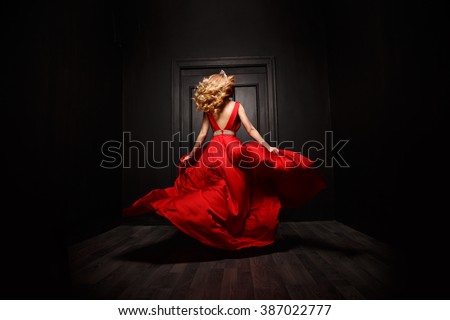 Elegant and sexy woman in the red evening fluttering dress is capture in move, running away from the ceremony (the 	
ball), the wooden door is on the background