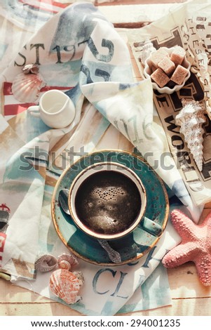 Cup of americano coffee set with seashells, milk and sugar on marine themed napkin. Holiday by tropical sea relaxation concept. Brightly sunlit setting, natural light. Toned photo.