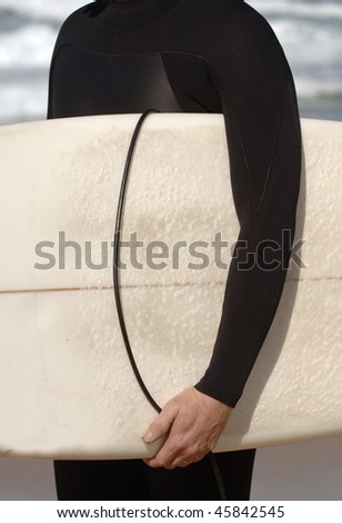 A detail of a man in a wet suit carrying a surfboard at the beach
