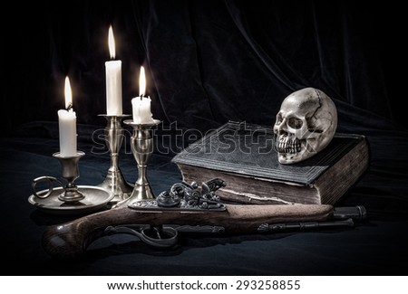 Still life of a skull on an old leather bound book, three lit candles in candlesticks and a replica 17th century flintlock pistol on a black curtain