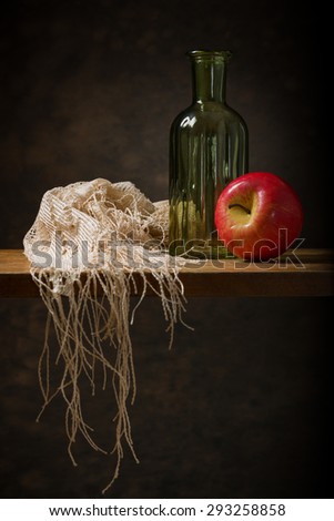 Still life of a woollen shawl, a red apple and a green bottle on a wooden shelf with a mottled brown background