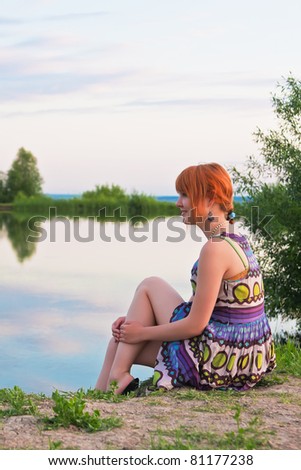 Beautiful ginger-haired woman in sundress sitting at the coast of pond