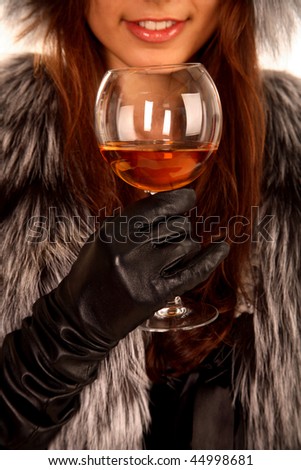 The girl dressed in a fur coat keeps in a hand a glass with an alcoholic drink and smiling