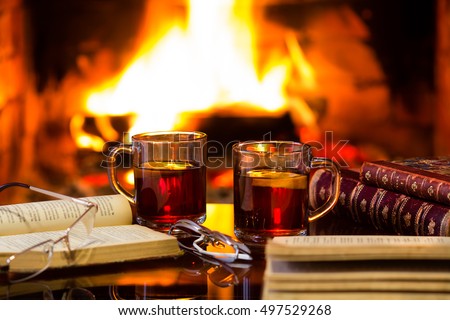 Two glass mugs of hot drink or alcoholic drink or mulled red wine and antique books in front of warm fireplace. Magical relaxed cozy atmosphere near fire. Autumn or winter concept
