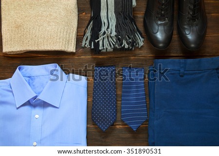 collection of business modern men\'s clothing: light brown sweater, scarf, black shoes, blue shirt, ties, blue pants for business travel on a dark wooden background.  Top view.