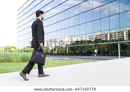 Anonymous businessman or worker in black suit with leather handbag or laptop case in his hand goes to work on the road to the entrance of a glass office building near green grass.