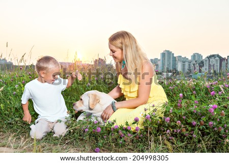 Happy family. Mother with son and dog playing on green meadow. Shape of city with sunset on background
