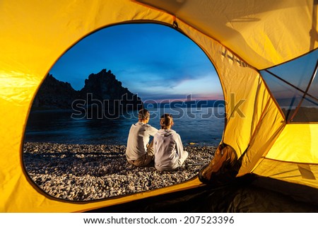 Couple sits near tent. Man and woman sit near tent at lake shore and look at sunset