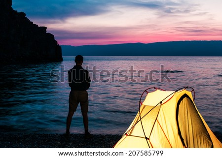 Man stand close to tent and lake shore and looking at sunset