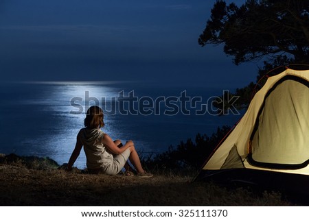 Woman near lit tent and looking at reflection of moon on the sea