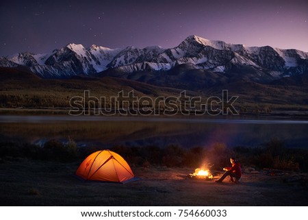 Night camping. Man tourists sitting in the illuminated tent near campfire under amazing sunset evening sky in a mountains area. Snow mountain in the background