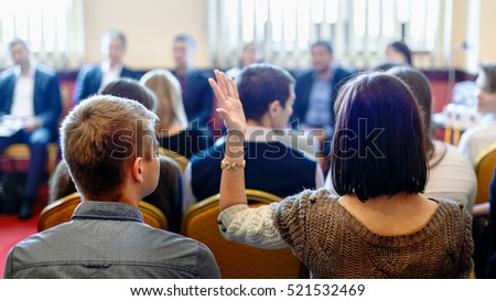 Young woman rear view raising hand up to make a speech at professor lecture in auditorium room