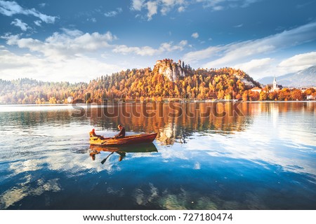 Romantic couple sailing by boat on Bled lake water in Slovenia at medieval fortress circled by colorful autumn forest background. Stunning fall scenery. Bled is famous and popular travel destination.