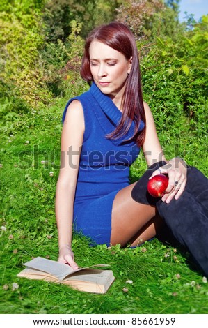 young woman is sitting on green grass reading a book and holding an apple in her hand