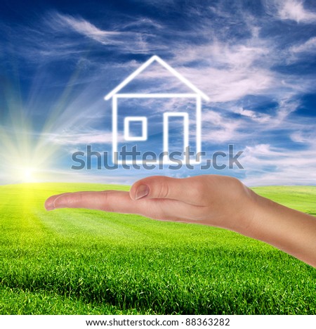 green field over cloudy blue sky with female hand holding white drawn house