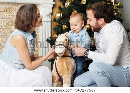 Happy smiling young family near the Christmas tree celebrating New Year. Mom dad and baby sitting on toy horse near the tree. Mother, father and their child on Christmas eve. Mom, dad and child.